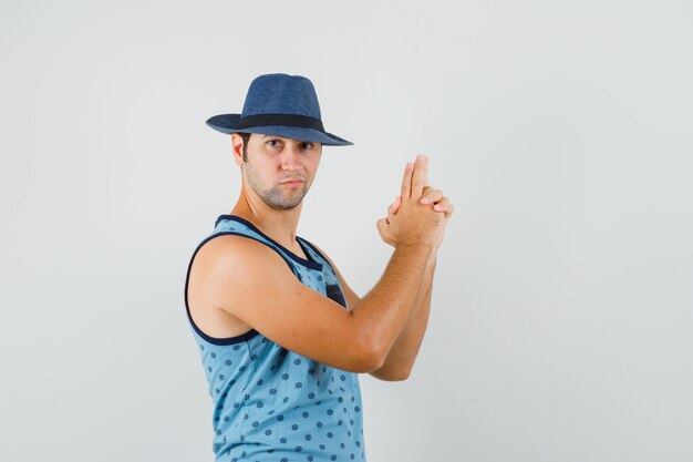 Joven mostrando gesto de pistola en camiseta azul, sombrero y mirando confiado. vista frontal.