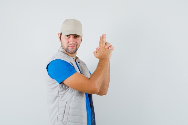 Joven mostrando gesto de pistola en camiseta, chaqueta, gorra y mirando confiado, vista frontal.