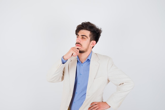 Joven mostrando gesto de silencio mientras sostiene la mano en la cintura con camisa azul y chaqueta de traje blanco y mirando pensativo