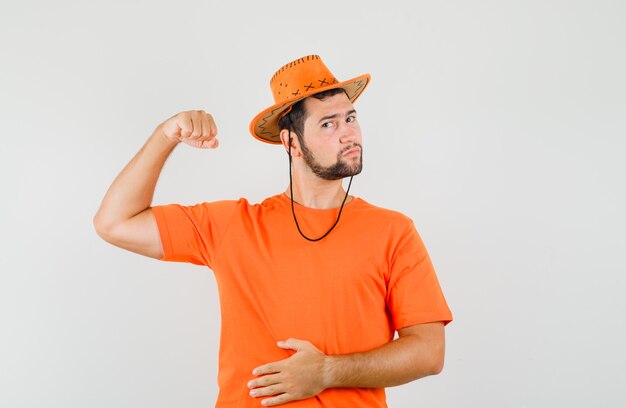 Joven mostrando los músculos del brazo en camiseta naranja, sombrero y mirando confiado. vista frontal.