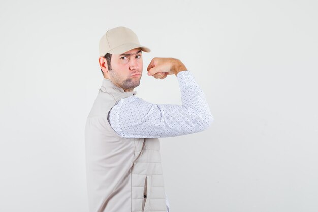 Joven mostrando músculos en chaqueta beige y gorra y mirando confiado, vista frontal.