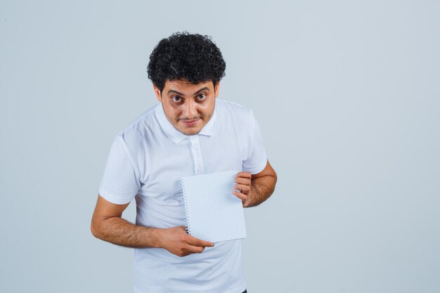 Joven mostrando portátil en camiseta blanca y jeans y mirando feliz, vista frontal.