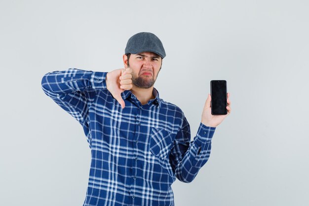 Joven mostrando el pulgar hacia abajo, sosteniendo el teléfono móvil en camisa, gorra y mirando insatisfecho. vista frontal.