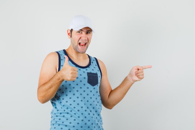 Joven mostrando el pulgar hacia arriba, apuntando a un lado en camiseta azul, gorra y mirando feliz. vista frontal.