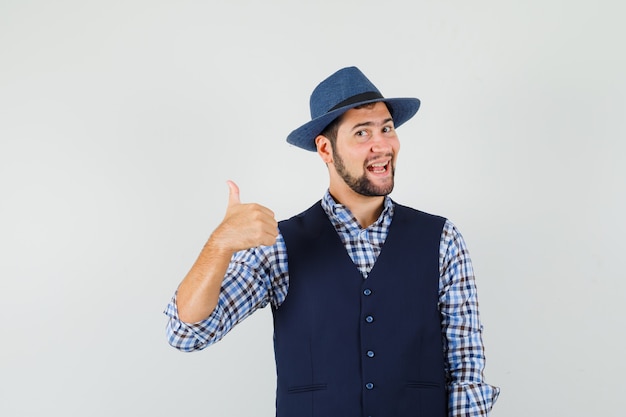 Foto gratuita joven mostrando el pulgar hacia arriba en camisa, chaleco, sombrero y mirando alegre.