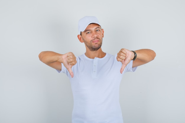 Joven mostrando los pulgares hacia abajo en camiseta blanca, gorra y mirando disgustado, vista frontal.