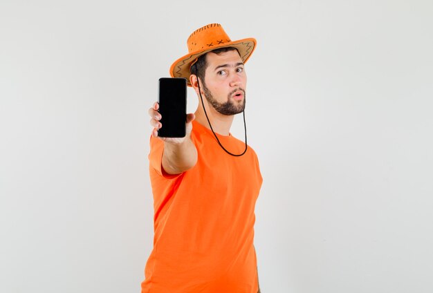 Joven mostrando teléfono móvil en camiseta naranja, sombrero, vista frontal.
