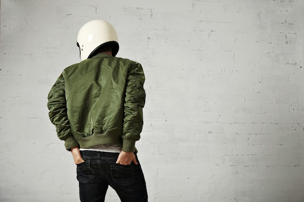 Joven motociclista en casco blanco y retrato de chaqueta verde desde atrás con las manos en los bolsillos traseros de sus jeans con paredes blancas.