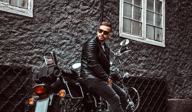 Foto gratuita un joven motociclista de moda con gafas de sol vestido con una chaqueta de cuero negro y jeans sentados en su motocicleta retro en la vieja calle europa.