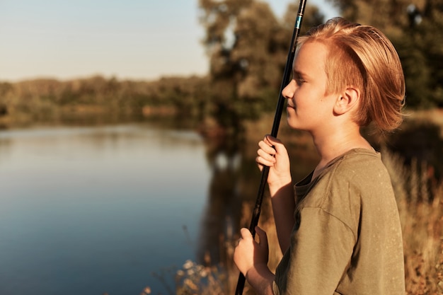 Joven muchacho rubio europeo pescando en un día soleado de verano, mirando en la distancia, disfrutando de su tiempo libre, vistiendo camiseta verde, de pie en la orilla del río cerca del agua.