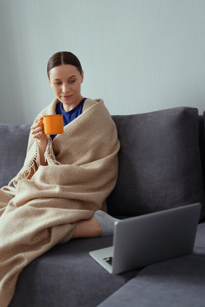 Joven mujer abrazada en una manta caliente con una computadora portátil y una taza de té