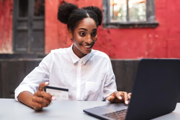 Joven mujer africana alegre con camisa blanca sosteniendo una tarjeta de crédito en la mano mientras trabaja felizmente en una laptop en el acogedor patio de la cafetería