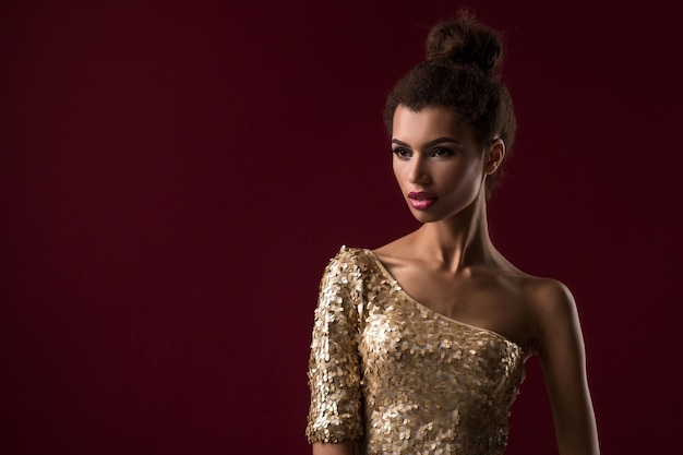 Joven mujer africana de moda con maquillaje, con un sexy vestido dorado. Modelo sobre un fondo de clarete en el estudio.