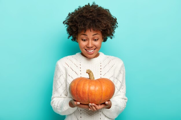 Joven mujer afro de pelo rizado con expresión feliz mira calabaza de otoño de cosecha propia, viste suéter blanco