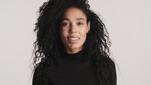 Foto gratuita joven mujer afroamericana bonita con el pelo oscuro y esponjoso vestido con un cuello de tortuga negro que parece feliz aislado de fondo blanco