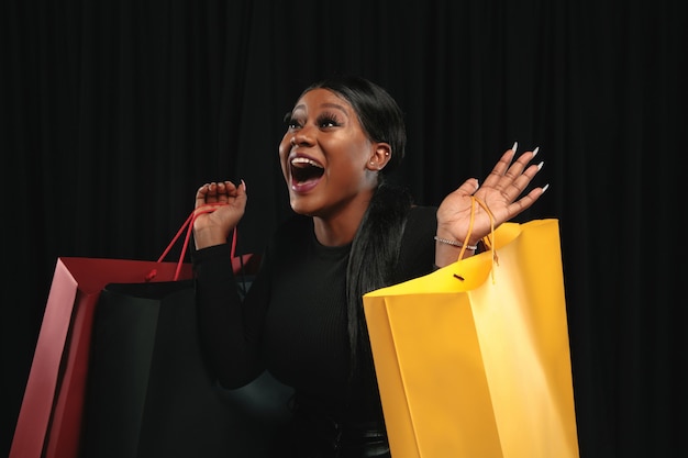 Joven mujer afroamericana de compras con paquetes de colores sobre fondo negro. Modelo femenino atractivo.