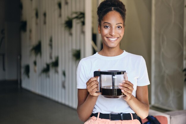 Joven mujer afroamericana muy sonriente con cafetera mirando alegremente a la cámara en un moderno espacio de coworking