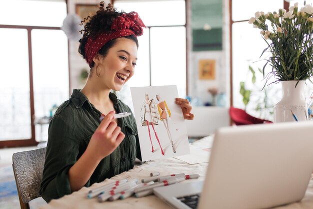 Joven mujer alegre con cabello oscuro y rizado sentada en la mesa mostrando felizmente un boceto de moda en una laptop mientras pasa tiempo en un taller moderno y acogedor con grandes ventanas