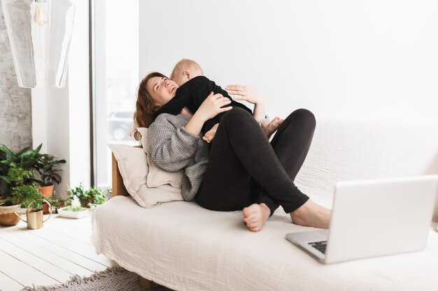 Joven mujer alegre en el sofá abrazando felizmente a su pequeño hijo Mamá y bebé pasando tiempo soñadoramente juntos con una computadora portátil en la acogedora sala de estar en casa