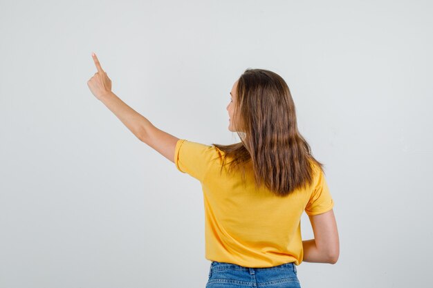 Joven mujer apuntando con el dedo hacia arriba y sonriendo en camiseta, pantalones cortos, vista posterior.