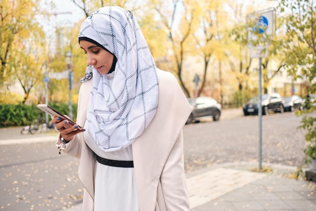 Joven mujer árabe atractiva en hijab usando intensamente el teléfono inteligente en la acogedora calle de la ciudad