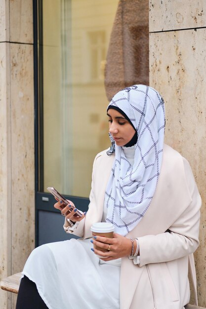 Joven mujer árabe atractiva con hiyab usando un teléfono inteligente mientras se sienta en la calle de la ciudad con café para llevar