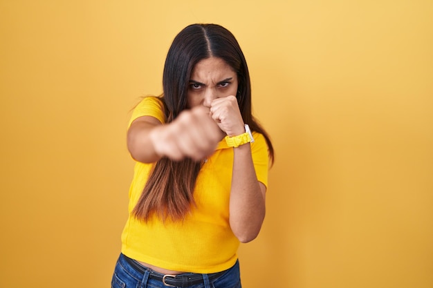 Foto gratuita joven mujer árabe de pie sobre fondo amarillo golpeando el puño para luchar contra la amenaza y la violencia de ataques agresivos y enojados