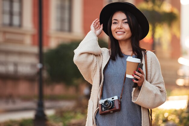 Joven mujer asiática encantadora en sombrero negro con café para llevar y teléfono móvil, mirando a un lado mientras camina en la calle al aire libre