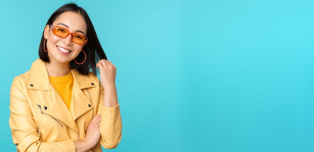 Joven mujer asiática con estilo en gafas de sol sonriendo jugando con su corte de pelo y luciendo feliz posando ag