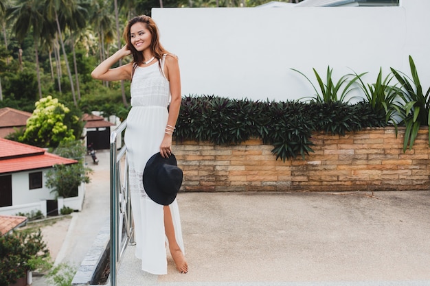 Joven mujer asiática con estilo en vestido blanco boho, estilo vintage, natural, sonriente, feliz, vacaciones tropicales, hotel, fondo de palmeras