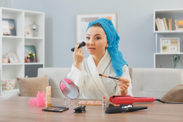 Joven mujer asiática feliz con una toalla en la cabeza sentada en el tocador en casa aplicando sombras de ojos y colorete haciendo rutina de maquillaje matutino concepto de belleza y cosmética facial