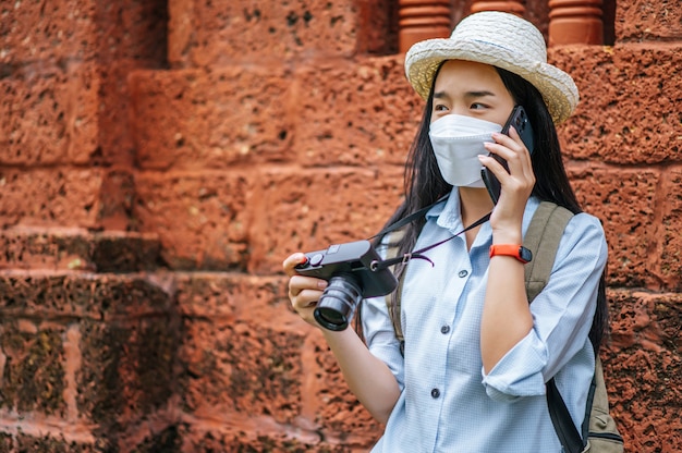 Joven mujer asiática mochilero con sombrero y máscara de protección mientras viaja en un sitio histórico, hablando con el teléfono inteligente y sosteniendo la cámara en la mano