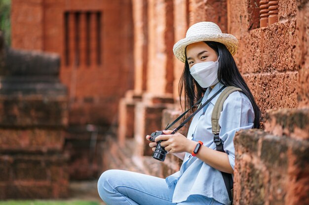 Joven mujer asiática mochilero con sombrero y máscara de protección mientras viaja en un sitio histórico