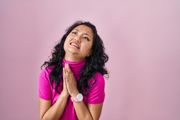 Foto gratuita joven mujer asiática de pie sobre un fondo rosado rogando y rezando con las manos juntas con expresión de esperanza en la cara muy emocional y preocupada. mendicidad.