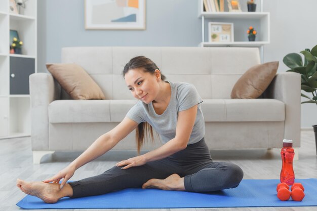 Joven mujer asiática saludable haciendo ejercicio en casa estirándose en la sala de estar sentada en una alfombra de yoga feliz y positiva