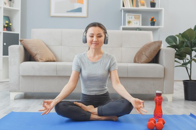 Joven mujer asiática sana con auriculares haciendo ejercicio en casa meditando en la sala de estar sentada en una alfombra de yoga haciendo gestos de meditación
