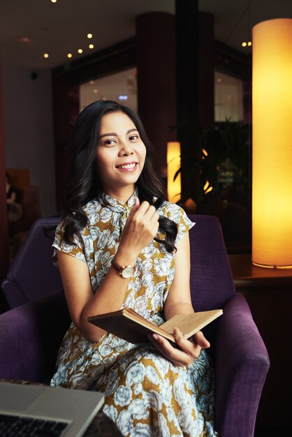 Joven mujer asiática sentada en un sillón en la cafetería con bolígrafo y diario