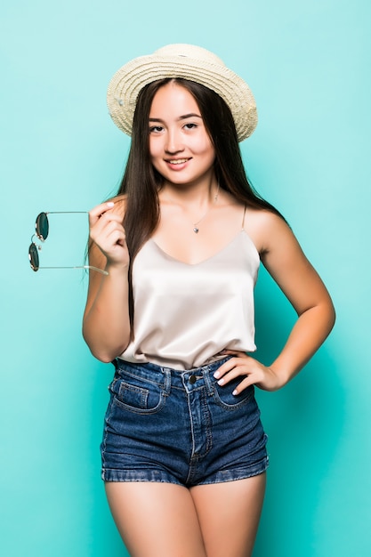 Foto gratuita joven mujer asiática en sombrero y gafas de sol sonriendo fondo turquesa