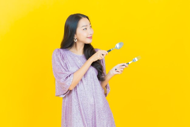 Joven mujer asiática sonriendo con cuchara y tenedor en amarillo