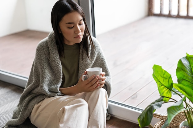Joven mujer asiática triste sentada en el piso cerca del balcón y mirando la taza de té pensativo