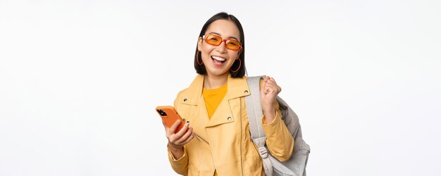 Joven mujer asiática viajera turista con gafas de sol sosteniendo mochila y teléfono móvil usando la aplicación en el teléfono inteligente de pie sobre fondo blanco