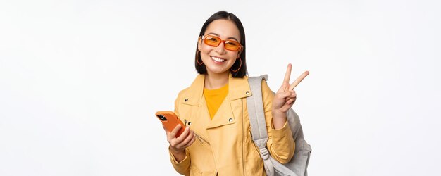 Joven mujer asiática viajera turista con gafas de sol sosteniendo mochila y teléfono móvil usando la aplicación en el teléfono inteligente de pie sobre fondo blanco