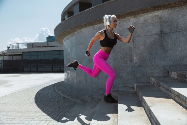Una joven mujer atlética en camisa y auriculares blancos trabajando escuchando música en la calle al aire libre. Corriendo escaleras arriba. Concepto de estilo de vida saludable, deporte, actividad, pérdida de peso.