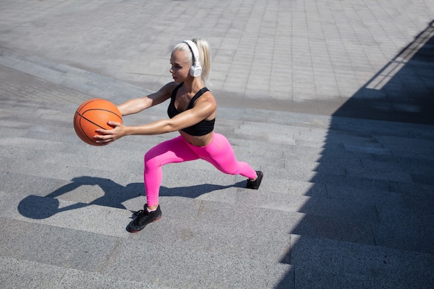 Una joven mujer atlética en camisa y auriculares blancos trabajando escuchando música en la calle al aire libre. Hacer estocadas con la pelota. Concepto de estilo de vida saludable, deporte, actividad, pérdida de peso.