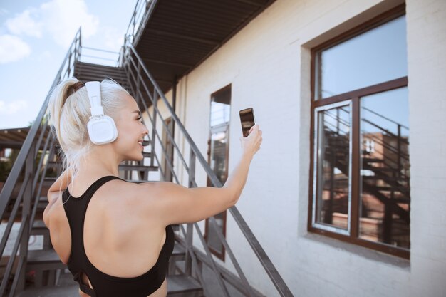 Una joven mujer atlética en camisa y auriculares blancos trabajando escuchando música en las escaleras al aire libre. Haciendo una selfie. Concepto de estilo de vida saludable, deporte, actividad, pérdida de peso.