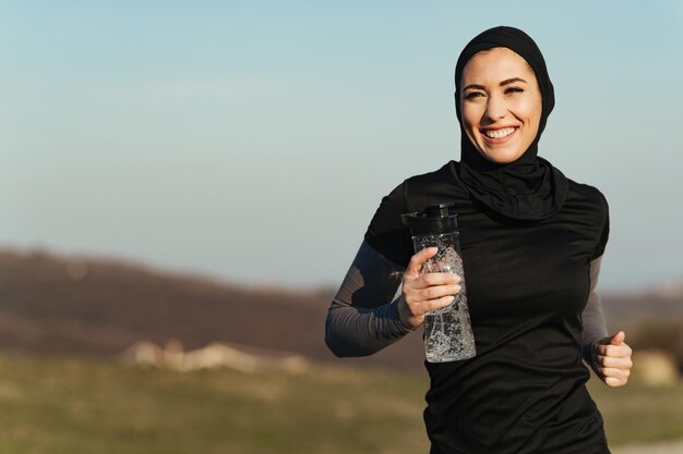 Joven mujer atlética feliz que lleva una botella de agua mientras trota en la naturaleza