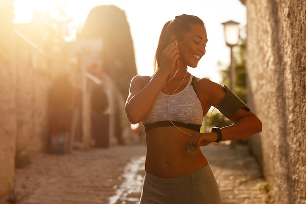 Joven mujer atlética feliz usando un teléfono inteligente en su banda de brazo y escuchando música mientras se prepara para hacer ejercicio en la ciudad