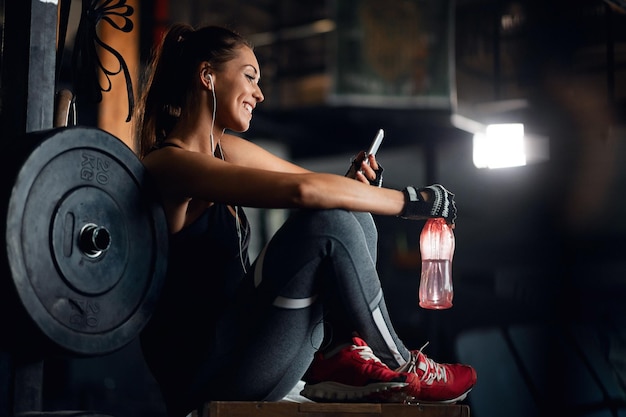 Joven mujer atlética sonriente relajándose en un descanso para tomar agua y leyendo un mensaje de texto en su teléfono inteligente en un gimnasio