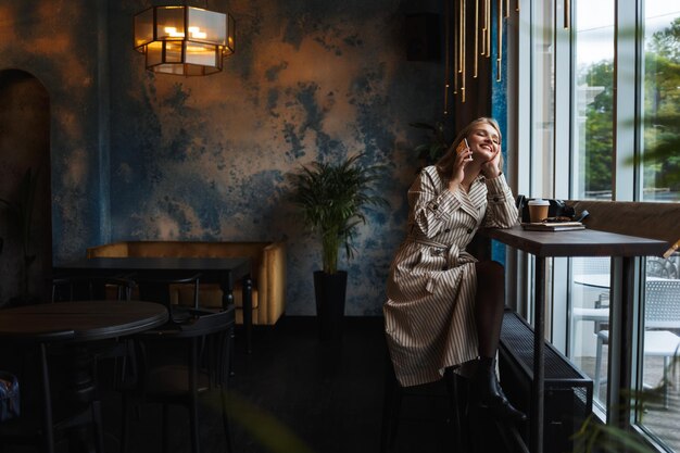 Joven mujer atractiva con gabardina a rayas hablando alegremente por teléfono celular sentada cerca de una gran ventana mientras pasa tiempo en un café moderno