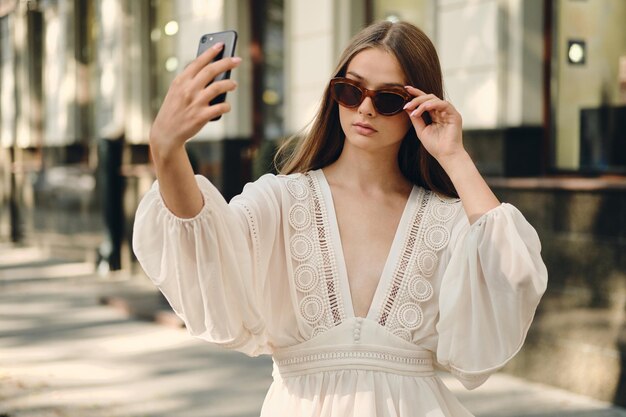 Joven mujer atractiva con gafas de sol y hermoso vestido blanco mirando cuidadosamente a un lado mientras toma una foto en el celular en la acogedora calle de la ciudad